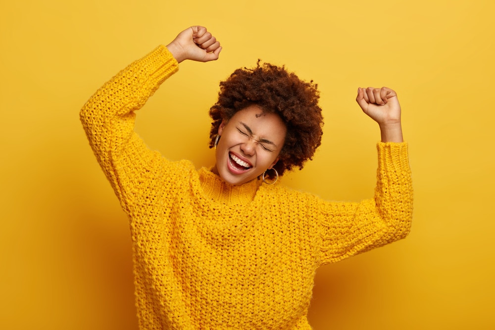 joyful afro woman raises arms tilts head dressed casual knitted jumper laughs from happiness celebrates victory isolated yellow