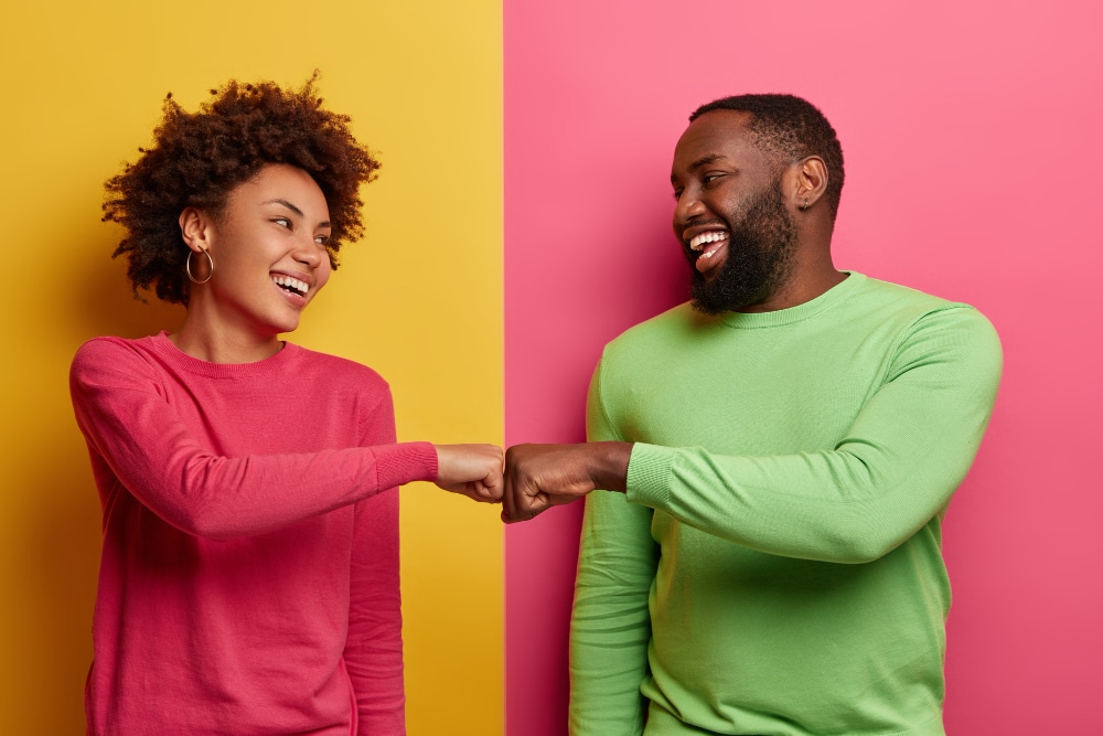 positive dark skinned young woman man bump fists agree be one team look happily each other celebrates completed task wear pink green clothes pose indoor have successful deal